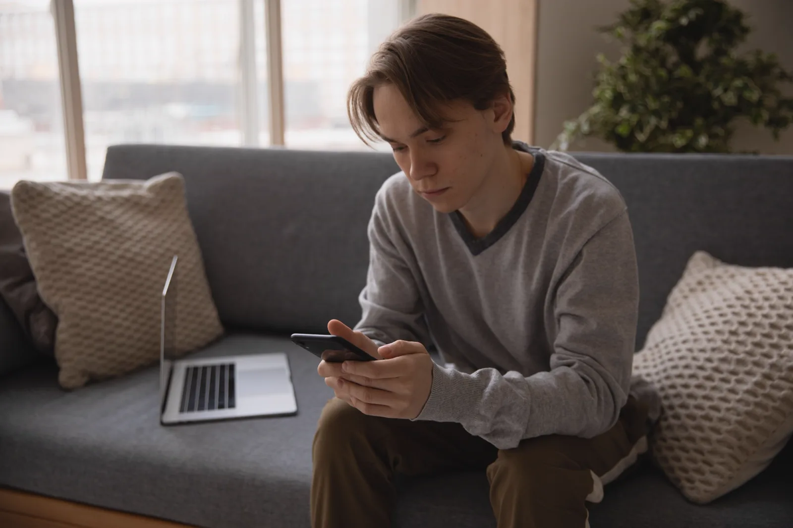 A person sitting on a sofa, looking down at their phone, with a laptop on the cushion beside them.